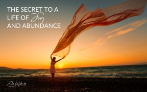 The Secret to a Life of Joy and Abundance | woman on a shore holding a long scarf caught up in the air with a backdrop of a sunset